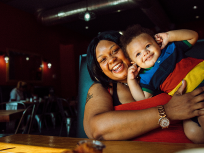 Robena Hill, a Black mother from Detroit, Mi, sits with her son at a booth at Cuzzo's. Toddler son is making silly faces at the camera.