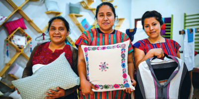 Three Indigenous women in Chiapas, Mexico, holding textile goods such as pillows and shirts.