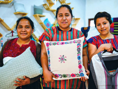 Three Indigenous women in Chiapas, Mexico, holding textile goods such as pillows and shirts.