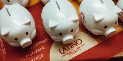White colored piggy banks on a table with the logo and text of the nonprofit, Latino Community Foundation of Colorado. The Organization is spearheading latinos making change in Colorado.