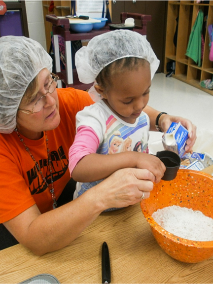 A child pouring milk into a baking bowl with the help of an early care and education provider in Waukon, IA. Photo courtesy of Northeast Iowa Food & Fitness Initiative.