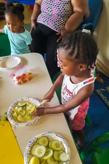 Two young girls help prepare their own snacks and meals by cutting fresh tomatoes and cucumbers. Photo courtesy of WECA and Mother and Daughter Child Nurturing Center in Milwaukee, WI.