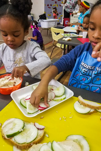 Two kids spread hummus and add cucumbers and radishes to their sandwiches for lunchtime. Photo courtesy of WECA and Mother and Daughter Child Nurturing Center in Milwaukee, WI.