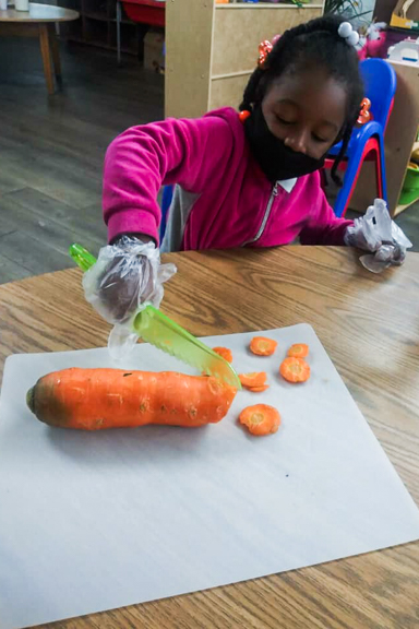 A young girl rolls up her sleeves to help cut slices of a freshly picked carrot for a midday snack. Photo courtesy of Little Ones Learning Center in Forest Park, GA.