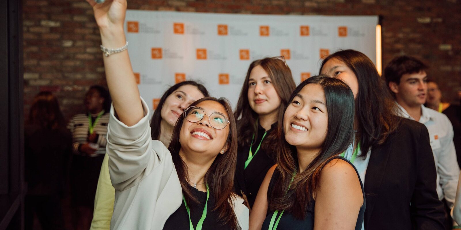 A group of smiling young professionals take a selfie together at the KFIO summer symposium, standing in front of a brick wall and a backdrop with event logos.