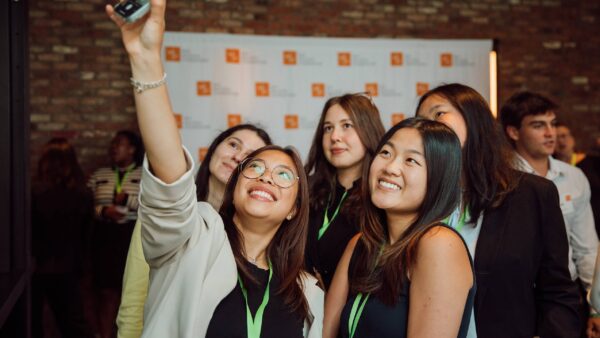 A group of smiling young professionals take a selfie together at the KFIO summer symposium, standing in front of a brick wall and a backdrop with event logos.
