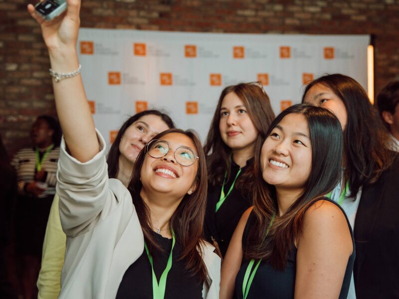 A group of smiling young professionals take a selfie together at the KFIO summer symposium, standing in front of a brick wall and a backdrop with event logos.