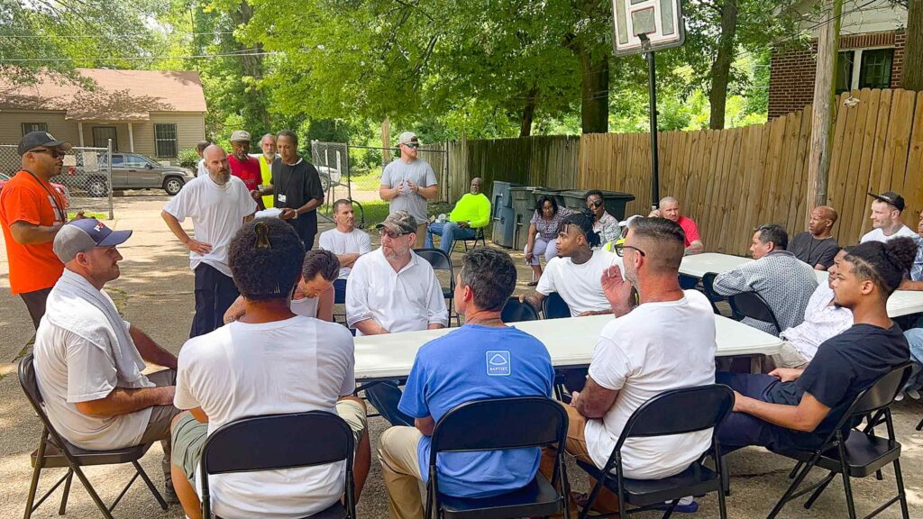 Outdoor gathering of people seated at tables listening to a speaker in a residential setting.