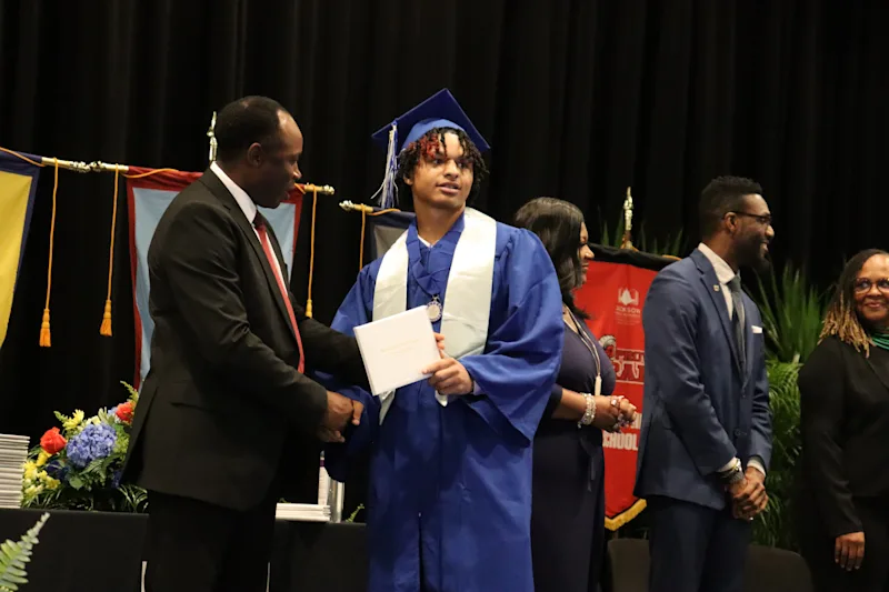 Graduate in blue cap and gown receiving a diploma and shaking hands on stage during a ceremony.