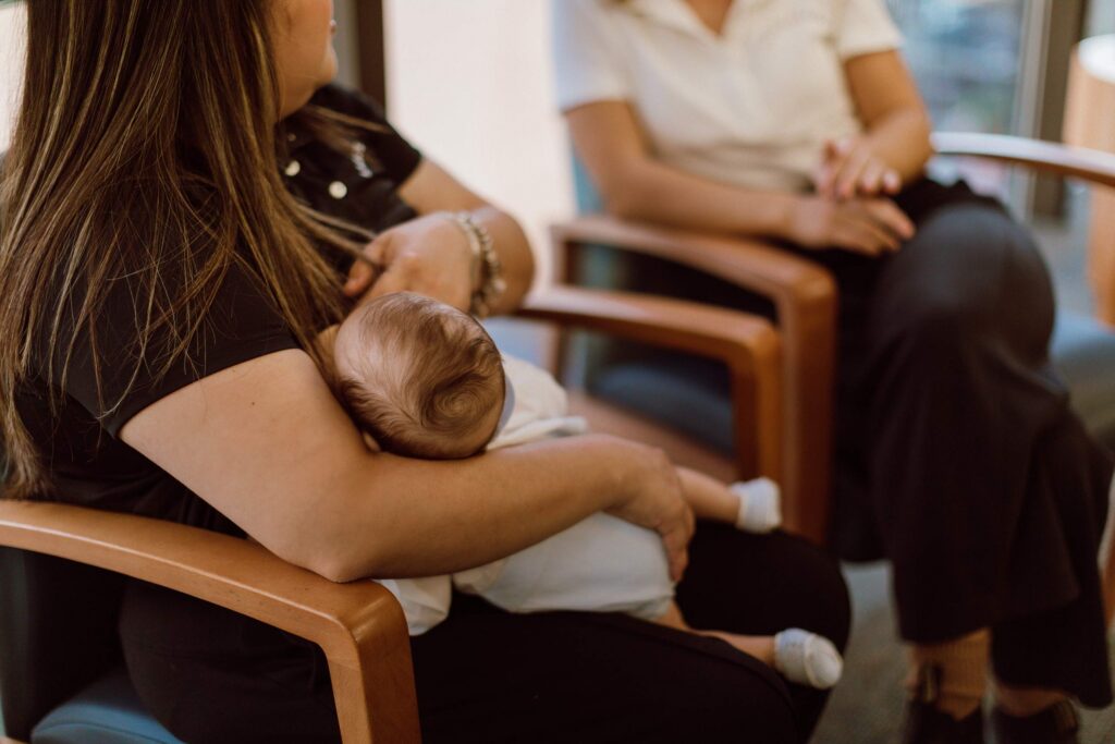 A woman holding a baby in her arms, breastfeeding, while sitting and talking with another person.