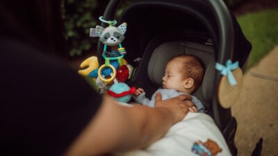A sleeping baby in a car seat stroller with colorful toys hanging above.