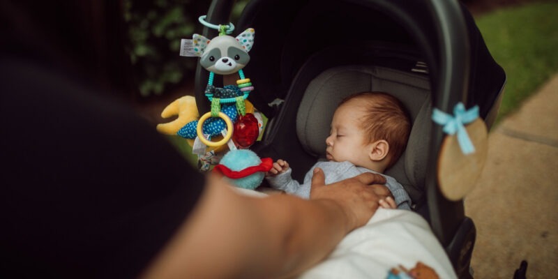 A sleeping baby in a car seat stroller with colorful toys hanging above.