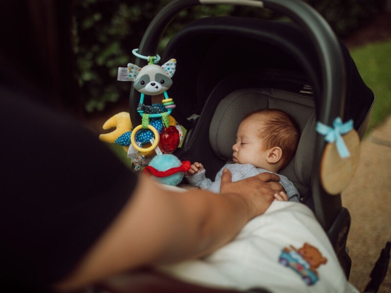 A sleeping baby in a car seat stroller with colorful toys hanging above.